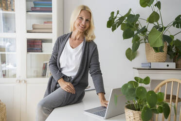 Smiling freelancer with laptop sitting on desk in home office - SVKF00507