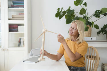 Smiling freelancer with wind turbine model sitting at desk in home office - SVKF00512