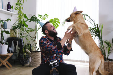 Glücklicher Mann mit Partyhut spielt mit Hund zu Hause - ASGF02670
