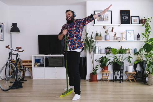 Mature man standing with broom singing in living room at home - ASGF02683