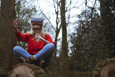 Young woman wearing VR glasses sitting cross-legged on tree trunk in forest - AMWF00310