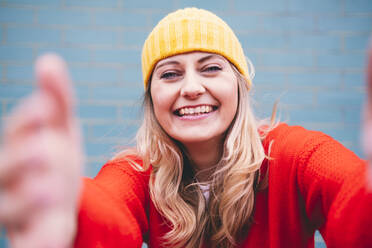 Glückliche junge Frau mit gelber Strickmütze vor einer Mauer - AMWF00315