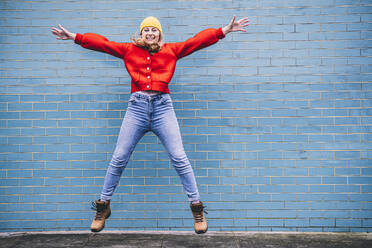 Carefree young woman jumping with arms outstretched in front of wall - AMWF00316