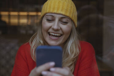 Glückliche Frau mit gelber Strickmütze, die zu Hause ein Mobiltelefon benutzt - AMWF00317