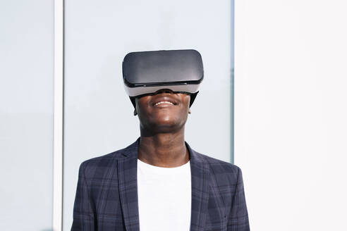 Lächelnder junger Mann mit VR-Brille vor einer Wand - AMWF00320