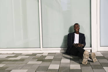Glücklicher junger Mann sitzt auf dem Boden vor einer Mauer - AMWF00340