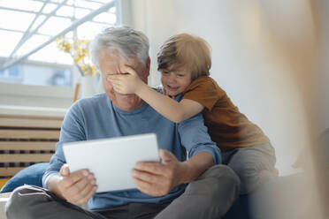 Boy covering eyes of grandfather with tablet PC at home - JOSEF12099