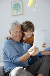 Smiling boy taking selfie with grandfather through tablet computer at home - JOSEF12115