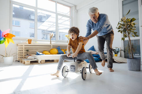 Cheerful grandfather pushing grandson sitting on toy car in living room - JOSEF12124