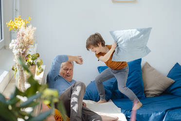 Happy boy playing pillow fight with grandfather on sofa at home - JOSEF12172