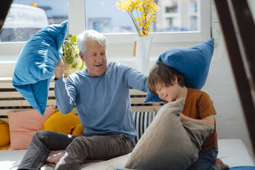 Senior man playing pillow fight with grandson in living room - JOSEF12174