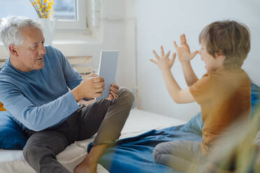 Senior man photographing playful grandson through tablet PC at home - JOSEF12187