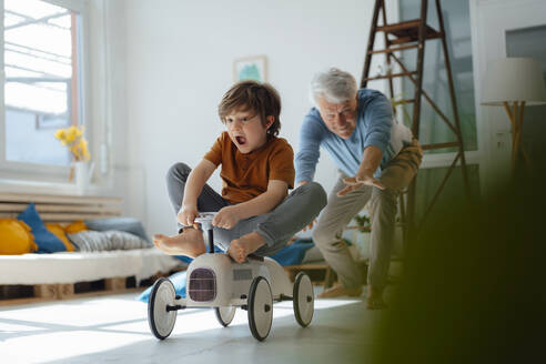 Cheerful grandfather playing with grandson sitting on toy car in living room - JOSEF12198