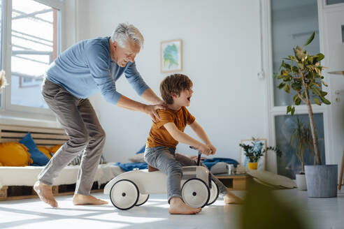 Verspielter älterer Mann, der seinen auf einem Spielzeugauto sitzenden Enkel im heimischen Wohnzimmer schiebt - JOSEF12203