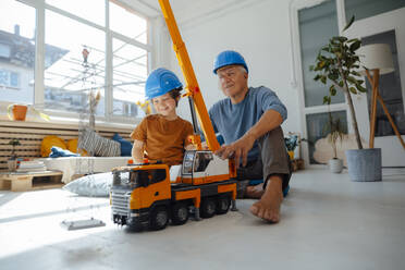 Lächelnder Enkel und Großvater in Schutzhelmen mit Spielzeugkran und Strommastmodell zu Hause - JOSEF12204