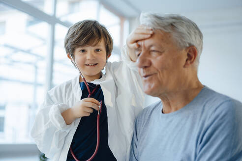 Smiling boy imitating as doctor checking grandfather's temperature at home - JOSEF12242