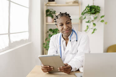 Smiling female doctor with tablet PC sitting at table in home office - JCZF01065