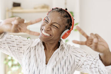 Cheerful woman wearing wireless headphones showing peace sign at home - JCZF01077