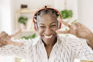 Fröhliche reife Frau mit drahtlosen Kopfhörern, die zu Hause eine Friedensgeste zeigt - JCZF01078