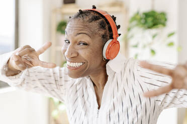 Happy woman wearing wireless headphones showing peace sign at home - JCZF01079