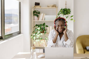 Smiling freelancer wearing wireless headphones looking at laptop - JCZF01081