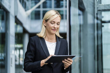 Smiling blond businesswoman using tablet PC in front of building - DIGF18644