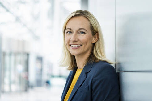 Smiling businesswoman with blond hair leaning on wall - DIGF18673