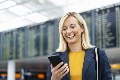 Happy businesswoman using mobile phone in front of departure board - DIGF18675