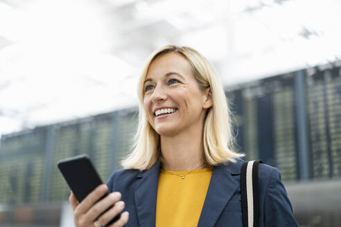 Happy businesswoman with mobile phone in front of arrival board - DIGF18676