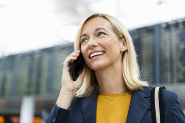 Smiling businesswoman with blond hair talking on phone at airport - DIGF18677