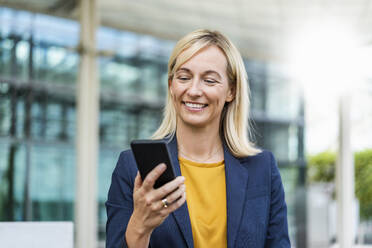 Happy businesswoman using smart phone standing in front of building - DIGF18678