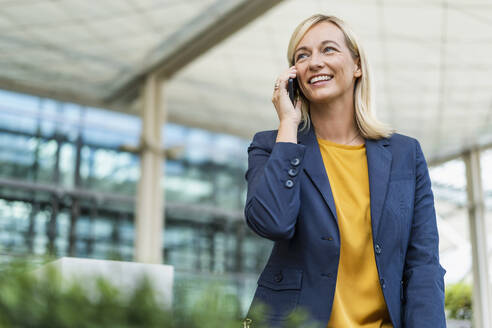 Smiling businesswoman talking on phone standing in front of building - DIGF18679