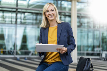 Smiling businesswoman with tablet PC sitting in front of building - DIGF18683