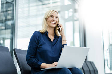 Lächelnde Geschäftsfrau mit Laptop, die in der Lobby telefoniert - DIGF18720