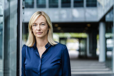 Mature businesswoman with blond hair leaning on glass wall - DIGF18727