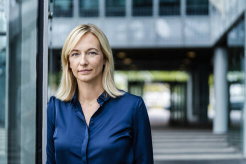 Mature businesswoman with blond hair leaning on glass wall - DIGF18727