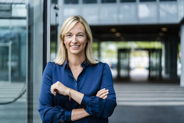 Smiling mature businesswoman standing by glass wall - DIGF18728