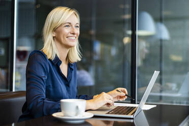 Smiling blond businesswoman with laptop sitting in cafe - DIGF18733