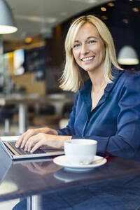 Glückliche Geschäftsfrau mit Laptop am Tisch im Café sitzend - DIGF18743