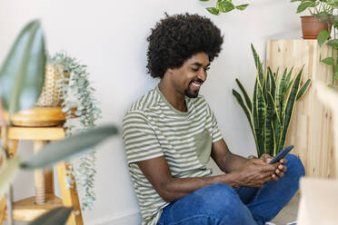Glücklicher Mann mit Afrofrisur, der zu Hause ein Smartphone benutzt - XLGF03022