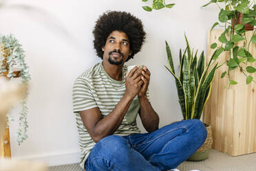 Afrikanischer Mann mit Kaffeetasse, der zu Hause vor einer Wand sitzt - XLGF03030