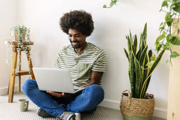 Glücklicher Mann, der einen Laptop benutzt und zu Hause vor einer Wand sitzt - XLGF03033