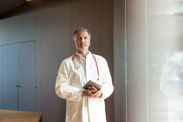 Confident senior doctor holding tablet PC near glass wall in hospital - JOSEF12261