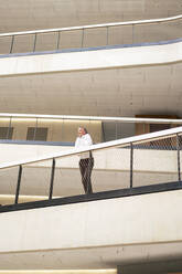 Businessman leaning on railing in corridor of office - JOSEF12308