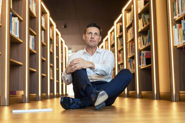 Nachdenklicher Mann sitzt in einer beleuchteten Bibliothek auf dem Boden - JOSEF12344