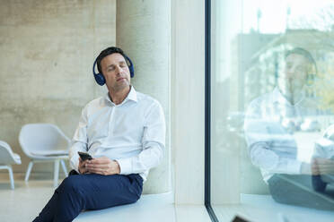 Geschäftsmann mit geschlossenen Augen hört Musik auf Kopfhörer sitzen in der Nähe von Fenster im Büro - JOSEF12351