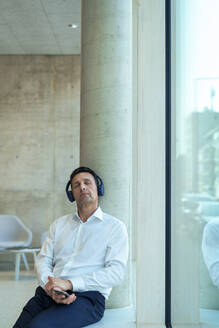 Geschäftsmann mit Kopfhörern sitzt an einer Säule in der Nähe des Fensters im Büro - JOSEF12352