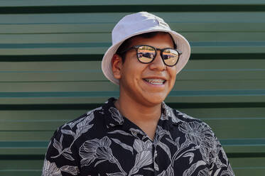 Happy young man with sunglasses and hat in front of wall - PGF01218