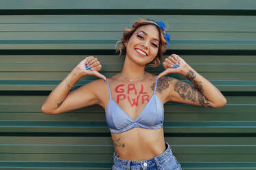 Glückliche junge Frau mit Girl-Power-Text auf der Brust vor einer grünen Wand - PGF01237