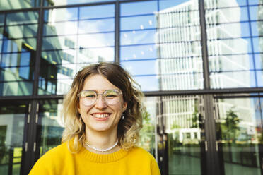 Happy young woman wearing eyeglasses standing in front of glass - IHF01139